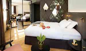 Hotellihuone, jossa on suuri sänky, seinäkoristeita ja peili, joka heijastaa huonetta. Matkalaukku seisoo vieressä, ja pöydällä on pieni kaktus. (Tekoälyn luoma teksti)