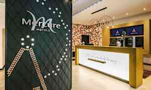 Mercure-hotellin vastaanottoalue, jossa näkyy yrityksen logo koristeellisella seinällä ja vastaanottotiski tekstillä "Let's make somebody happy". (Tekoälyn luoma teksti)