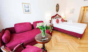 Hotellihuone, jossa on punainen sohva, pyöreä pöytä kukkakimpulla ja punaisin yksityiskohdin varustettu sänky. (Tekoälyn luoma teksti)