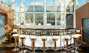 Cocktail-baari, jossa on korkeat tuolit ikkunaseinän edessä, näkymä värikkäällä katolla varustettuun rakennukseen. (Tekoälyn luoma teksti)