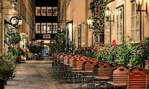 Viehättävä kuja, jossa ravintoloiden ulkopuolella on tuoleja ja pöytiä, ympäröitynä kukkivilla pensailla ja lyhdyillä. (Tekoälyn luoma teksti)