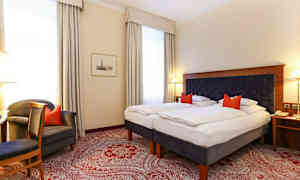 Hotellihuone, jossa on kaksi yhden hengen vuodetta, tummasta kankaasta valmistettu sängynpääty ja punaiset koristetyynyt. Huoneessa on valkoiset seinät, suuret ikkunat vaaleilla verhoilla ja punainen kuvioitu matto. (Tekoälyn luoma teksti)