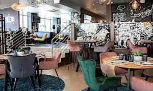 Moderni ravintolan sisustus, jossa on värikkäitä tuoleja pöytien ympärillä, joille on katettu lautasia ja laseja. Seinät on koristeltu taiteellisilla kuvioilla ja teksteillä. (Tekoälyn luoma teksti)