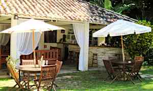 Ulkoilmaympäristö, jossa puisia pöytiä ja tuoleja sekä aurinkovarjoja, rakennuksen vieressä, jossa tiilikatto ja sisäverhot. (Tekoälyn luoma teksti)