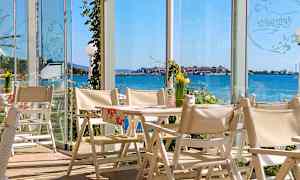 Näköala ravintolasta, jossa on valkoisia tuoleja ja pöytiä sekä lasiseinät merelle ja rannikkokaupunkiin. (Tekoälyn luoma teksti)