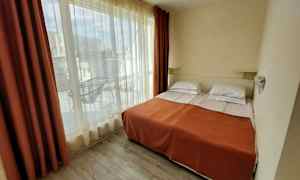 Hotellihuone, jossa kaksi erillistä vuodetta oranssin peitteen alla ja suuret ikkunat, jotka avautuvat parvekkeelle. (Tekoälyn luoma teksti)