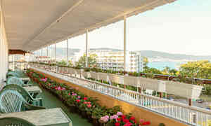 Terassi, jossa vihreitä muovituoleja ja pöytiä, kukkaruukkujen ympäröimänä ja näkymä hotellille ja merelle. (Tekoälyn luoma teksti)