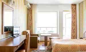 Hotellihuone, jossa on kaksi sänkyä, puinen työpöytä, jolla on TV, pieni sohvanurkkaus sohvapöydällä ja parveke taustalla. (Tekoälyn luoma teksti)