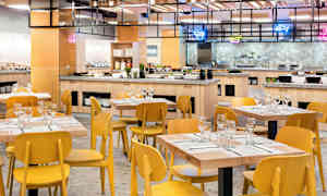 Kuva ruokasalista, jossa on puupöytiä ja keltaisia tuoleja, sekä taustalla buffet. (Tekoälyn luoma teksti)