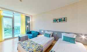 Hotellihuone, jossa on kaksi yhden hengen sänkyä, sininen nojatuoli ja pöytä. Huoneessa on vaaleat seinät ja suuri ikkuna, jossa on keltainen verho ja parveke. (Tekoälyn luoma teksti)