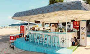 Rantabaari, jossa siniset tuolit ja katto, sijaitsee lähellä hiekkarantaa. (Tekoälyn luoma teksti)