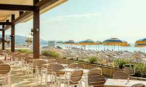 Ulkoterassi, jossa pöytiä ja tuoleja, edustalla ranta aurinkotuoleineen ja varjoineen. (Tekoälyn luoma teksti)