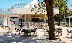 Ulkobaari, jossa on pöytiä ja tuoleja puiden alla, vaalealla sisustuksella ja puukoristeilla. (Tekoälyn luoma teksti)