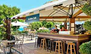 Kahvila, jossa on ulkona pöytiä ja tuoleja Sunshine Caféssa, taustalla vehreyttä ja merinäköalaa. (Tekoälyn luoma teksti)