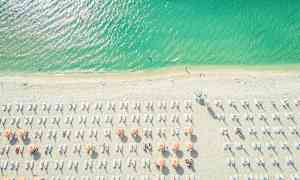 Ilmakuva rannasta, jossa on aurinkovarjoja ja aurinkotuoleja. Meri on kauniin sinivihreä. (Tekoälyn luoma teksti)