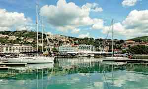 Useita purjeveneitä on kiinnitetty satamaan, taustalla kaupunki ja kukkuloita. (Tekoälyn luoma teksti)