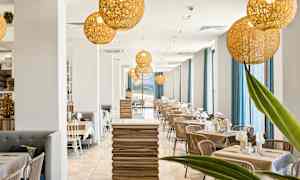 Moderni ravintola, joka on sisustettu vaaleilla puupöydillä, tuoleilla ja suurilla ikkunoilla, joissa on siniset verhot. Katosta roikkuu koristeellisia valaistusarmatuureja. (Tekoälyn luoma teksti)