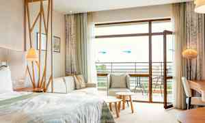 Hotellihuone, jossa on sänky, oleskelualue, pöytä ja avoin parvekkeen ovi. (Tekoälyn luoma teksti)