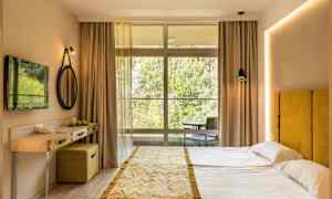 Hotellihuone, jossa on parisänky, keltainen sängynpääty, pieni kirjoituspöytä ja suuri ikkunaseinä kohti luontoa. (Tekoälyn luoma teksti)