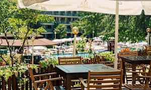 Ulkoilmapaikka, jossa puisia pöytiä ja tuoleja aurinkovarjon alla, puutarhan ja uima-allasalueen lähellä. (Tekoälyn luoma teksti)