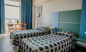 Kaksi yhden hengen sänkyä huoneessa, jossa on sininen ja harmaa sisustus. Pieni pöytä ja kaksi tuolia sijaitsevat suuren ikkunan ääressä. (Tekoälyn luoma teksti)