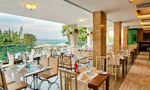 Ravintola, jossa pöytiä ja vaaleasävyisiä tuoleja, näkymä merelle ja ympärillä puita. (Tekoälyn luoma teksti)