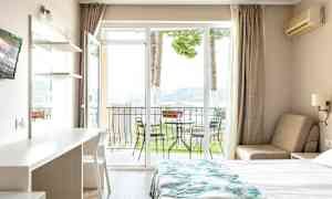 Hotellihuone, jossa on sänky, pöytä ja tuoli, sekä parveke ulkokalusteineen ja näkymä luontoon. (Tekoälyn luoma teksti)
