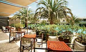 Ulkotarjoilutila, jossa pöytiä ja tuoleja lähellä uima-allasta, jota ympäröivät palmut. (Tekoälyn luoma teksti)