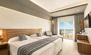 Hotellihuone, jossa on kaksi sänkyä ja oleskelutila, lasioven vieressä, joka johtaa parvekkeelle näkymällä. (Tekoälyn luoma teksti)