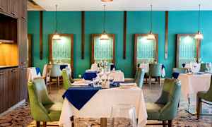 Tyylikäs ravintolasali, jossa on katettuja pöytiä ja vihreitä tuoleja, ympäröitynä turkoosilla seinillä ja koristeellisilla varjostimilla. (Tekoälyn luoma teksti)