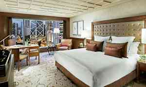 Hotellihuone, jossa on suuri sänky, nojatuoli ja työpöytä. Ikkunasta näkyy kaupunkinäkymä korkeine rakennuksineen. (Tekoälyn luoma teksti)