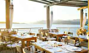 Ulkoilmaravintola, jossa pöytiä veden äärellä, näkymä merelle ja vuoristoon auringonlaskun aikaan. (Tekoälyn luoma teksti)