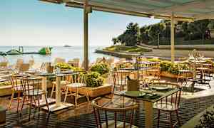 Ulkoterassi meren rannalla, jossa on pöytiä ja tuoleja katon alla, ympärillä kasveja ja taustalla ranta. (Tekoälyn luoma teksti)