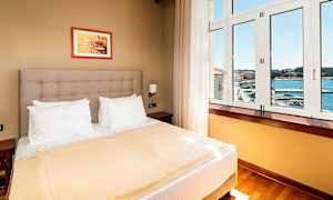 Hotellihuone, jossa on parivuode, valkoiset tyynyt, beige päiväpeite ja iso ikkuna, josta näkyy merimaisema. (Tekoälyn luoma teksti)