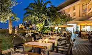 Iltaisin kuva ulkoilmassa sijaitsevasta ravintolasta, jossa on valaistuja pöytiä ja ympäröivänä palmuja sekä kasvillisuutta. (Tekoälyn luoma teksti)