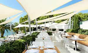 Ulkotila, jossa on useita pöytiä ja tuoleja jännitettyjen aurinkovarjojen alla lähellä vettä. (Tekoälyn luoma teksti)