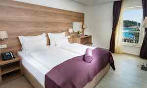 Hotellihuone, jossa on parivuode, valkoiset lakanat ja violetti päiväpeitto. Huoneessa on kirjoituspöytä tuolilla ja peili. Ikkunasta on näkymä merelle. (Tekoälyn luoma teksti)