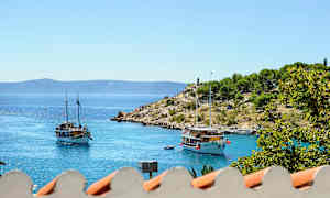 Kuvassa on kaksi purjevenettä tyynellä ja sinisellä merellä. Taustalla näkyy pieni niemi vehreyden ympäröimänä. Etualalla näkyy osia katosta. (Tekoälyn luoma teksti)