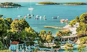 Näkymä uima-allasalueelle, jossa on aurinkotuoleja vehreyden ympäröimänä. Taustalla näkyy ranta, veneitä ja lahti pienine saarineen. (Tekoälyn luoma teksti)