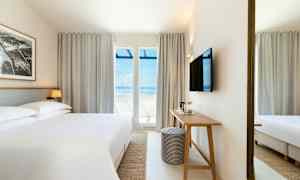 Hotellihuone, jossa iso sänky, kirjoituspöytä ja seinälle kiinnitetty televisio. Suuret ikkunat avautuvat merelle. (Tekoälyn luoma teksti)