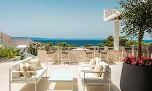 Terassi, jossa on istuimet ja pöytä sekä näkymä merelle. Ympäröiviä kasveja ja puita kirkkaan taivaan alla. (Tekoälyn luoma teksti)