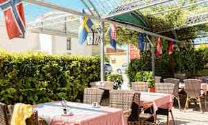 Ulkoterassi, jossa pöytiä, tuoleja ja lippuja vehreyden keskellä. (Tekoälyn luoma teksti)