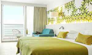 Hotellihuone, jossa on vihreät vuodevaatteet, seinäkoristeet ja istuinryhmä ikkunan vieressä parvekkeelle. (Tekoälyn luoma teksti)