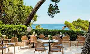 Kuva ulkoterassista, jossa on pöytiä ja tuoleja, ympärillä vehreyttä ja merinäköala. (Tekoälyn luoma teksti)