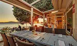 Ravintolapöydät sinivalkoruutuisilla pöytäliinoilla merenrannalla terassilla auringonlaskun aikaan. (Tekoälyn luoma teksti)