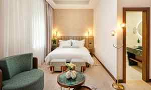 Tyylikäs hotellihuone, jossa on pedattu sänky, nojatuoli ja pieniä pöytiä. Kylpyhuone näkyy avonaisesta ovesta. (Tekoälyn luoma teksti)