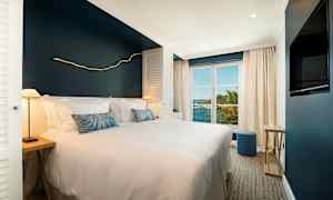 Hotellihuone, jossa on leveä vuode, valkoiset vuodevaatteet ja tummansininen seinä, suuret ikkunat päästävät luonnonvaloa ja tarjoavat näkymän trooppiseen kasvillisuuteen ja merelle. (Tekoälyn luoma teksti)