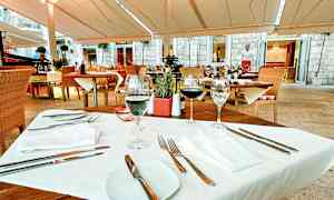 Ulkoravintolan pöytä, jossa puiset kalusteet ja valkoiset tekstiilit, katettuna vieraita varten. (Tekoälyn luoma teksti)