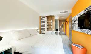 Moderni hotellihuone, jossa on suuri sänky valkoisilla vuodevaatteilla, oranssit ja valkoiset seinät, seinälle asennettu televisio ja työpöytä. (Tekoälyn luoma teksti)