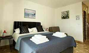 Hotellihuone, jossa sänky, sininen päiväpeite, kaksi tyynyä ja päälle taitellut pyyhkeet. Seinillä on tauluja, ja sängyn vieressä on yöpöytä, jossa on punainen lamppu. (Tekoälyn luoma teksti)